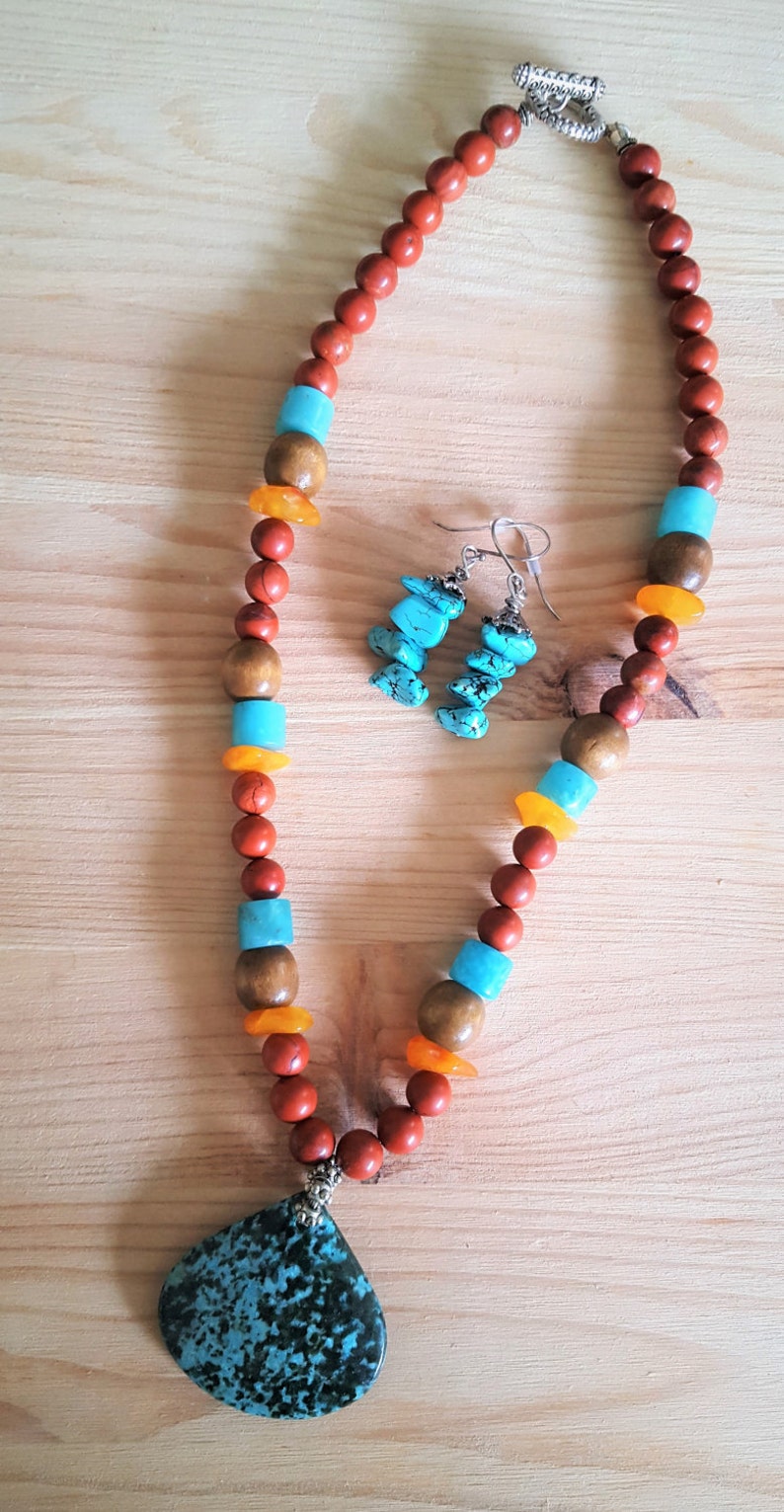Puede incluir: Un collar de cuentas turquesa y naranja con un gran colgante turquesa y pendientes a juego. El collar est&aacute; hecho con cuentas de madera y tiene un cierre plateado.