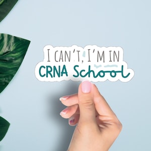 May include: A white sticker with the text "I can't, I'm in CRNA School" in teal and gray lettering. The sticker is held by a hand against a light blue background, with green leaves in the corner.