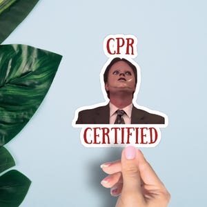 May include: A sticker featuring a character from a TV show, with the words "CPR CERTIFIED" in red. The sticker is held against a light blue background with green leaves.