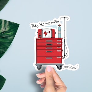 Peut inclure: Un autocollant représentant un chariot médical rouge avec des tiroirs, un support IV et le texte "They See me rollin'". Le chariot a un moniteur avec un symbole d'éclair. L'autocollant est sur un fond bleu clair.