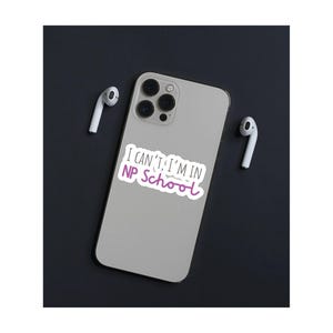 May include: A white iPhone with a sticker that says "I can't, I'm in NP School" in black and purple text. The phone is on a black surface with two white wireless earbuds.