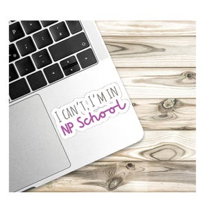 May include: A white laptop with a sticker that says "I can't, I'm in NP school". The sticker is on the top right corner of the laptop. The laptop is on a wooden surface.