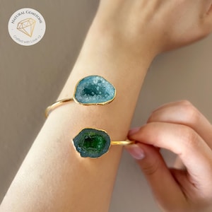 Op de afbeelding: Een goudkleurige manchetarmband met twee natuurlijke edelsteenaccenten. Eén accent is een lichtblauwe geode, de andere een donkergroene geode. De armband wordt om de arm van een persoon gedragen.
