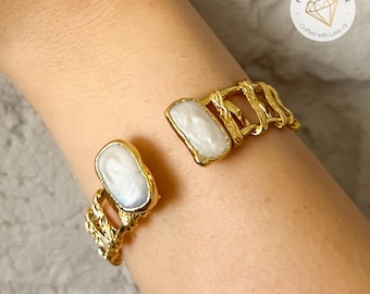 Puede incluir: Brazalete de puño dorado con dos piedras preciosas blancas grandes de forma irregular. El brazalete tiene un diseño de metal retorcido y texturizado. Las piedras preciosas están engastadas en un bisel dorado. El brazalete es abierto y ajustable.