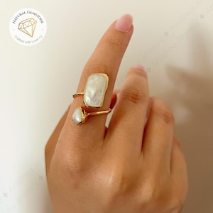 Puede incluir: Un anillo dorado con dos piedras blancas de forma irregular. Una es rectangular, la otra ovalada. El anillo está en un dedo. El anillo es de Anathea Jewelry. La imagen también incluye el texto "NATURAL GEMSTONE Crafted with Love <3".