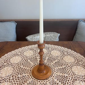 May include: A tall, white candle sits in a turned wooden candlestick on a crocheted lace tablecloth. The candlestick is made of light brown wood and has a round base. The tablecloth is cream-colored.