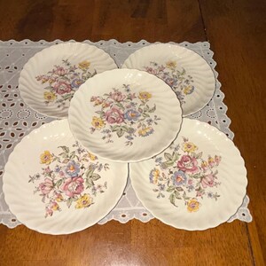 May include: A set of five off-white ceramic plates with a scalloped, swirl-patterned edge. Each plate features a central floral arrangement with pink, yellow, and blue flowers surrounded by green leaves and stems, arranged in a symmetrical pattern.
