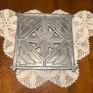 May include: A square, silver-colored metal trivet with an ornate, raised design. The center features a starburst pattern, surrounded by geometric shapes and leaf-like details. The trivet is placed on a white, lace doily, set against a wooden surface.