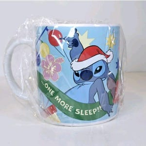 May include: A light blue ceramic mug featuring the character Stitch from Lilo & Stitch wearing a Santa hat. The mug has a white handle and is wrapped in clear plastic. The mug has the text "...ONE MORE SLEEP!!" on a green banner.