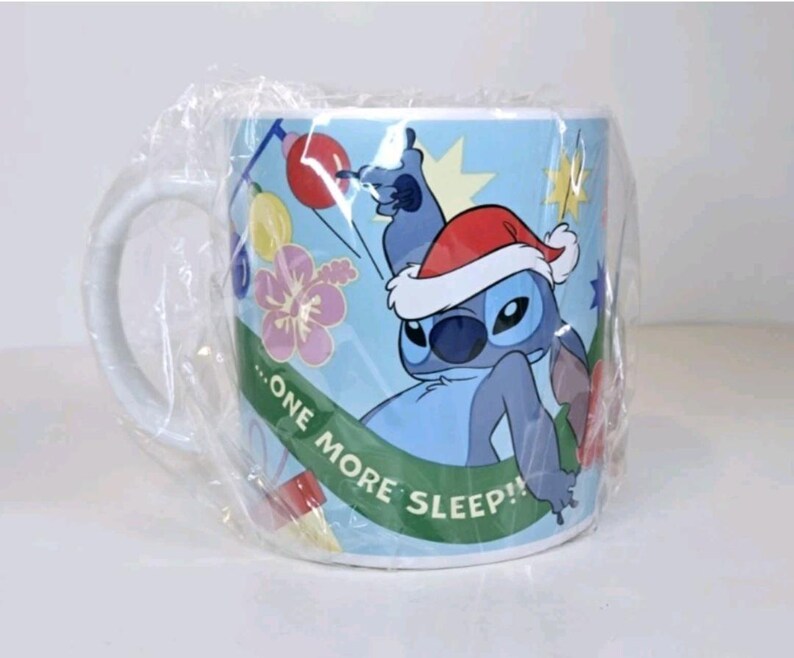 May include: A light blue ceramic mug featuring the character Stitch from Lilo & Stitch wearing a Santa hat. The mug has a white handle and is wrapped in clear plastic. The mug has the text "...ONE MORE SLEEP!!" on a green banner.