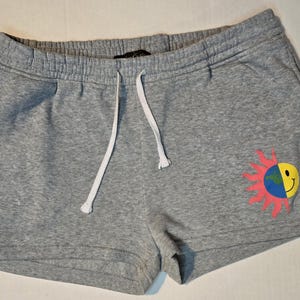 May include: Gray sweat shorts with a white drawstring and two front pockets. The shorts feature a colorful graphic of a smiling sun and earth design on the lower right leg.