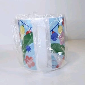 May include: White ceramic mug in clear wrapping. The mug has a light blue background with floral and ornament designs. The mug has a white handle and the text "...ONE" is visible.