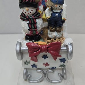以下が含まれることがあります： 装飾的なそりに乗った雪だるまを特徴とするお祝いのクリスマスフィギュア。雪だるまは帽子とスカーフを着用し、そりは赤いリボンと星のアクセントで飾られています。ベースは白で、雪に似ています。