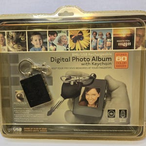May include: A digital photo album with a keychain, in a clear plastic case. The black, rectangular album displays a photo on its small screen. The packaging reads "Digital Photo Album with Keychain" and "Stores 60 Colour Images".