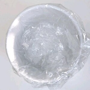 May include: A white, circular bowl is partially covered by transparent plastic wrap. The bowl seems to be empty and is set against a plain white background. The plastic wrap is crumpled and fills the inside of the bowl.