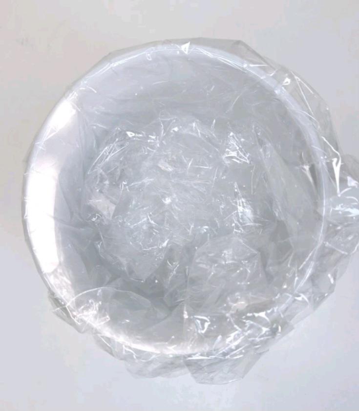 May include: A white, circular bowl is partially covered by transparent plastic wrap. The bowl seems to be empty and is set against a plain white background. The plastic wrap is crumpled and fills the inside of the bowl.