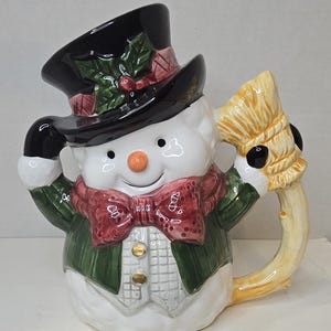 Könnte beinhalten: Eine Keramik-Schneemann-Tasse mit schwarzem Zylinder, grüner Jacke und roter Fliege. Die Tasse hat einen Besengriff und ein lächelndes Gesicht mit einer Karottennase. Eine festliche Weihnachtsdekoration.