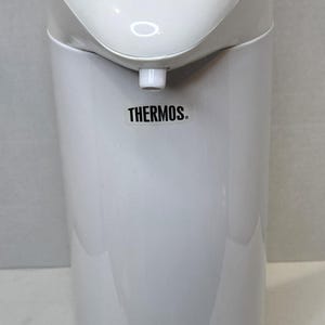 May include: White Thermos water boiler with a curved top and a cylindrical base. The boiler has a spout, a control panel with "CLOSE" and "OPEN" indicators, and the brand name "THERMOS" printed on the front.