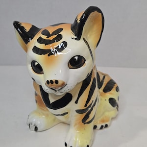 May include: A ceramic tiger cub figurine with a cream-colored face and body, adorned with black stripes and markings. The ears are tan with black tips, and the eyes are large and dark. The cub is in a seated position.