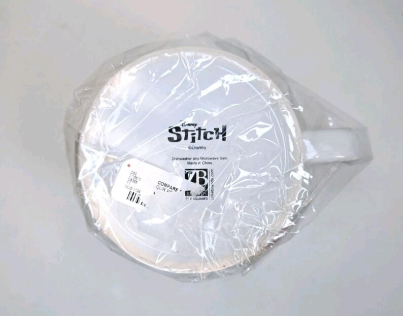 May include: A white ceramic mug with a handle, encased in clear plastic. The mug displays the word "Stitch" and the Disney logo. The mug is dishwasher and microwave safe.