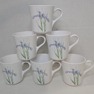 May include: A set of seven white ceramic mugs. Each mug features a watercolor-style illustration of a purple iris flower with green stems and leaves. The mugs have a curved handle and a simple, elegant design.