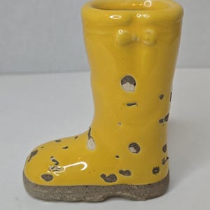 Op de afbeelding: Een kleine, gele keramische laars met een gespikkeld ontwerp. De laars heeft een afgeronde neus en een klein strikdetail bij de bovenkant. De basis van de laars is neutraal grijs van kleur. Dit decoratieve item is waarschijnlijk een kleine vaas of plantenbak.