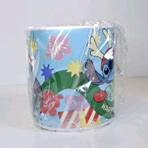 May include: A cylindrical mug wrapped in clear plastic. The mug has a light blue background with cartoon illustrations of Stitch, flowers, and stars. The word "HAPPY" is visible on the mug's side.