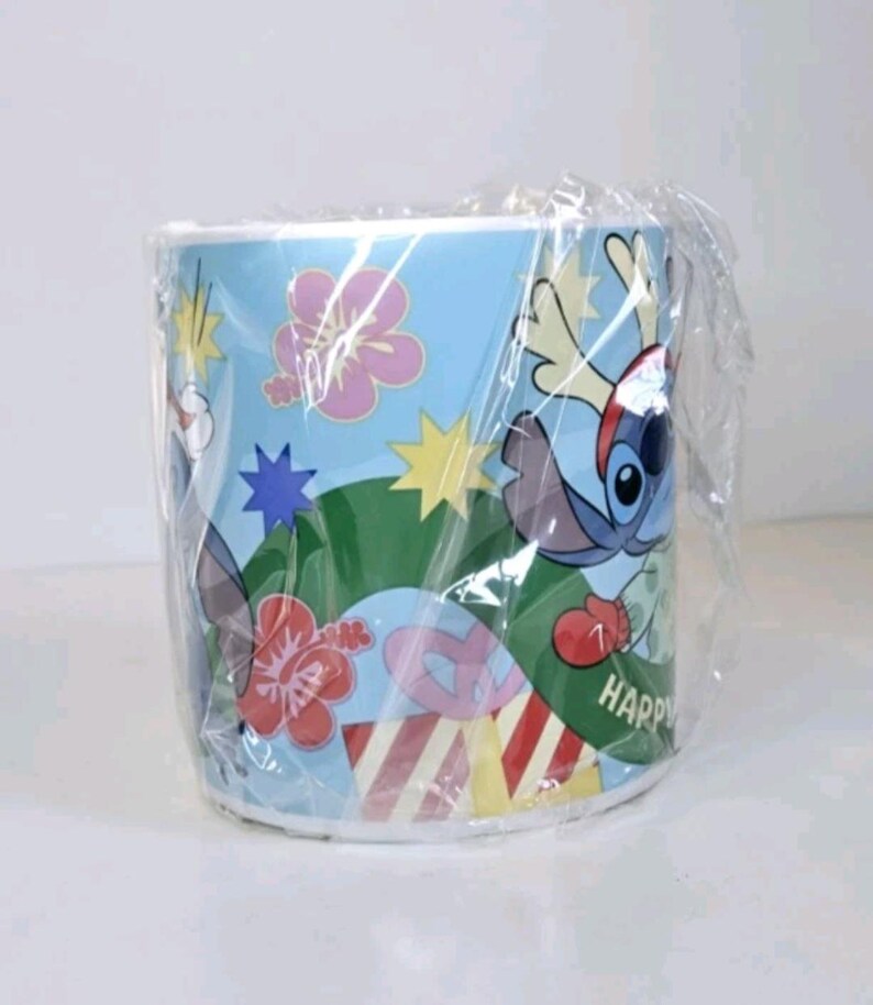 May include: A cylindrical mug wrapped in clear plastic. The mug has a light blue background with cartoon illustrations of Stitch, flowers, and stars. The word "HAPPY" is visible on the mug's side.