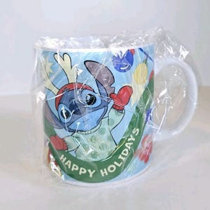 May include: A white ceramic mug featuring Stitch from Lilo & Stitch in a festive holiday design. Stitch is wearing a red hat and mittens, with the words "HAPPY HOLIDAYS" on a green banner. The mug is wrapped in clear plastic.
