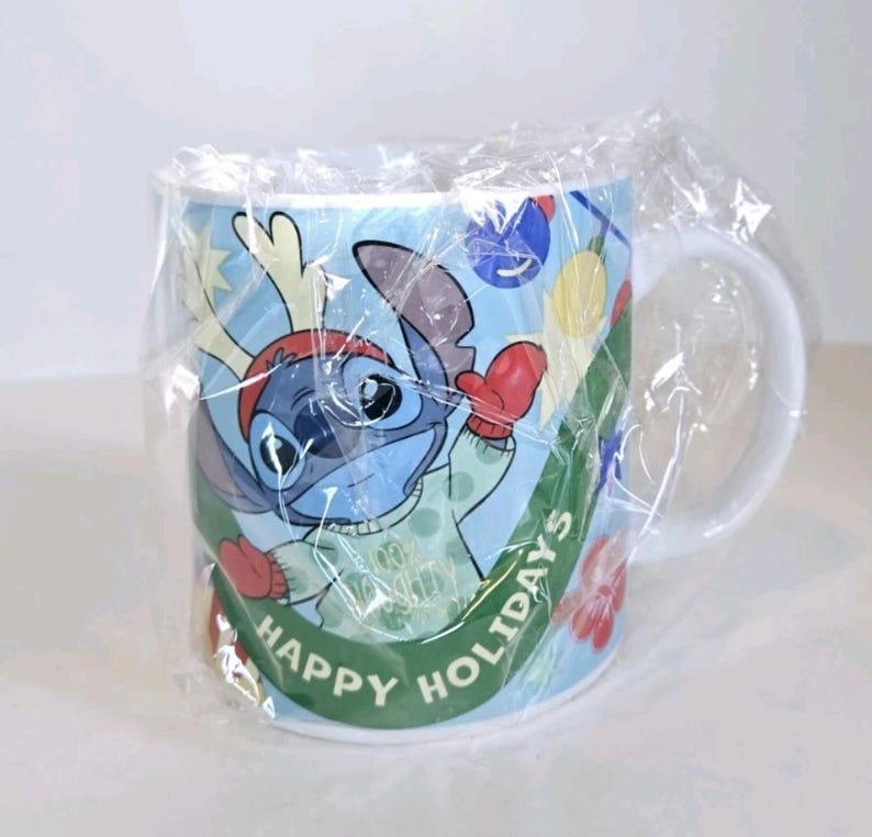 May include: A white ceramic mug featuring Stitch from Lilo & Stitch in a festive holiday design. Stitch is wearing a red hat and mittens, with the words "HAPPY HOLIDAYS" on a green banner. The mug is wrapped in clear plastic.