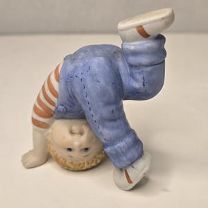 Può includere: Statuina in ceramica di un bambino in verticale. Il bambino indossa una tuta blu, una camicia a righe arancioni e bianche e scarpe bianche con dettagli arancioni. La statuina ha i capelli biondi e gli occhi blu.