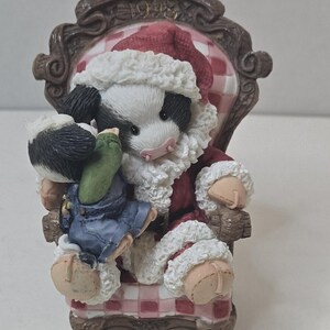 Könnte beinhalten: Figur einer Kuh als Weihnachtsmann, die in einem dekorativen Stuhl sitzt. Die Kuh trägt einen roten Weihnachtsmannanzug mit weißer Verzierung und einen passenden Hut. Der Stuhl hat ein rot-weißes Karomuster.