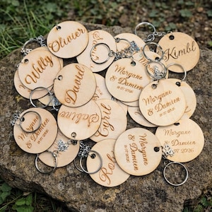Peut inclure: Plusieurs porte-clés ronds en bois clair, personnalisés avec des noms et des dates gravés. Chaque porte-clé est équipé d'un anneau argenté. On peut lire des noms comme "Olivier" et "David" sur certains d'entre eux.