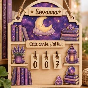 Può includere: Una decorazione da parete in legno con sfondo stellato viola, una luna crescente e il nome Sovanna. Presenta ripiani con libri, un vaso di lavanda, una lanterna, una sfera di cristallo e tre targhette in legno appese con il numero 007.