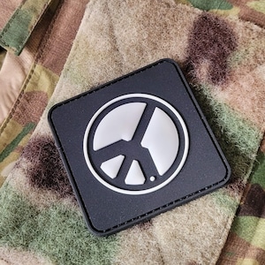 Könnte beinhalten: Ein schwarzer Patch mit einem weißen Friedenssymbol in der Mitte. Der Patch ist an einem Tarnstoff befestigt.