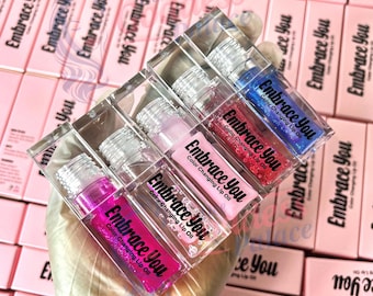 Puede incluir: Cuatro botellas de vidrio transparente con aceite labial de colores. Las botellas están etiquetadas con "Embrace You Color Changing Lip Oil" en rosa, rojo, azul y morado.