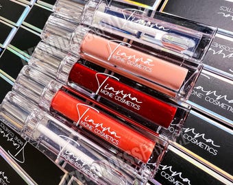 Può includere: Una collezione di tubetti di lucidalabbra in contenitori rettangolari trasparenti con tappi argentati, recanti la scritta Tianna Mone Cosmetics. I tubetti contengono tonalità trasparenti, nude, rosso scuro, arancione brillante e glitterate.