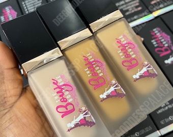 May include: Three bottles of The Bouje Boutique Pure Matte foundation in varying shades. Each bottle has a black cap with gold trim and a frosted glass body. The bottles are held in a hand, with boxes of the product visible in the background.