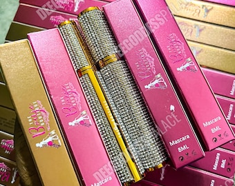 Puede incluir: Una colección de tubos y cajas de máscara de pestañas. Los tubos de máscara están adornados con pedrería y detalles dorados. Las cajas son rosas y doradas, con el texto "The Boutique" y "Mascara 8ML".