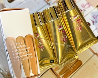 Puede incluir: Tubos de base de maquillaje líquida dorados con cajas a juego. Los tubos tienen una tapa dorada y el texto "Bouje The Boutique" y "Liquid Foundation SPF 15". Las cajas muestran muestras de base y características del producto.