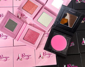 Puede incluir: Surtido de paletas de sombras de ojos rosas y negras con espejos. Cada paleta tiene un tono de sombra de ojos diferente y el texto "The Bouje Boutique" está impreso en la parte delantera de cada paleta.