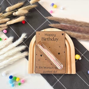 May include: A wooden rainbow arch with a brown card that says "Happy Birthday" and a small glass tube filled with colourful birthday candles. The card also says "Ein Wunsch, den du pustest du!"
