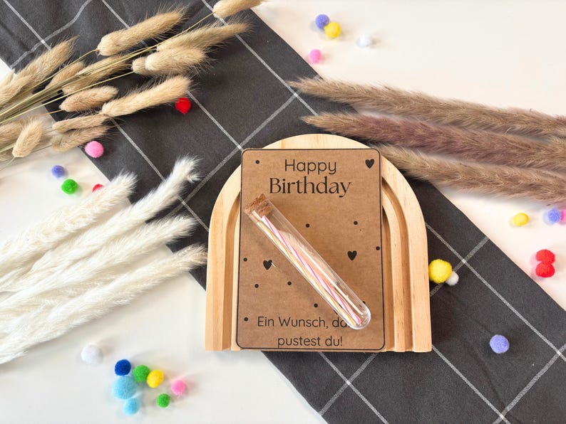 May include: A wooden rainbow arch with a brown card that says "Happy Birthday" and a small glass tube filled with colourful birthday candles. The card also says "Ein Wunsch, den du pustest du!"