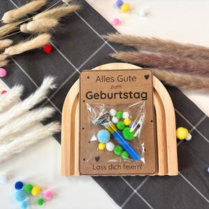 May include: A wooden rainbow arch with a card that says "Alles Gute zum Geburtstag. Lass dich feiern!"  The card has a small plastic bag with a blue party blower, green and white pom-poms, and a blue and white striped ribbon.