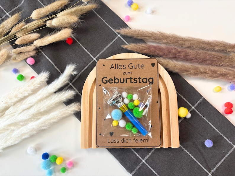 May include: A wooden rainbow arch with a card that says "Alles Gute zum Geburtstag. Lass dich feiern!"  The card has a small plastic bag with a blue party blower, green and white pom-poms, and a blue and white striped ribbon.