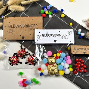 Puede incluir: Una variedad de pequeños regalos, que incluyen caramelos con forma de mariquita, un oso de papel dorado y pompones de colores, se exhiben sobre una superficie a cuadros. Los regalos están empaquetados en bolsas transparentes y acompañados de tarjetas con el texto "GLÜCKSBRINGER für dich."
