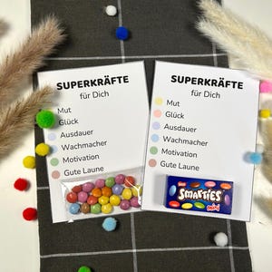 May include: Two white cards with the German text "SUPERKRÄFTE für Dich" and a list of words. A small bag of colorful candies and a box of "Smarties mini" are also present. The background includes decorative elements.