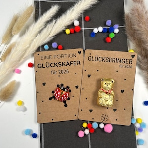 Peut inclure: Deux petites cartes avec le texte "EINE PORTION GLÜCKSKÄFER für 2026" et "GLÜCKSBRINGER für 2026". L'une des cartes représente une coccinelle, l'autre un ours en papier doré. Les cartes sont sur un tissu à carreaux gris avec des pompons colorés et des fleurs séchées.