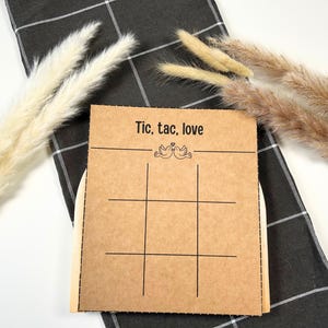 Könnte beinhalten: Ein braunes Tic-Tac-Toe-Spielkarten mit den Worten "Tic, tac, love" und einer Taubengrafik. Die Spielkarte liegt auf einem schwarz-weiß karierten Tuch, mit dekorativen getrockneten Pflanzen im Hintergrund.