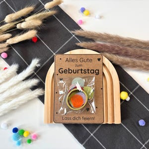 May include: A wooden rainbow arch with a card that says "Alles Gute zum Geburtstag. Lass dich feiern!" and a small bag of colourful confetti inside.